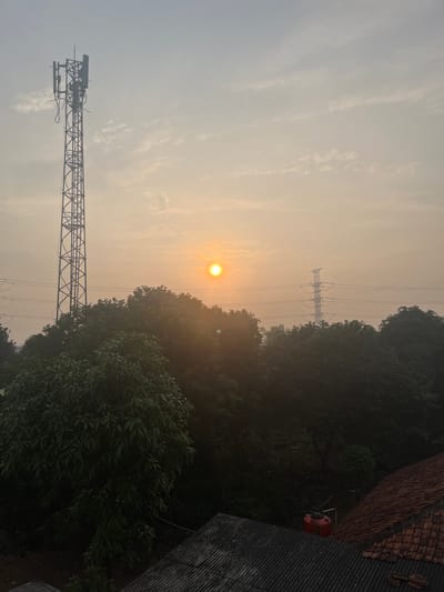 The image captures a hazy sunrise over a semi-urban landscape in Pasir Gadung, Indonesia. A tall telecommunications tower stands prominently on the left side of the frame, its metallic structure silhouetted against the pale sky. Power lines stretch across the middle ground, supported by pylons. Dense foliage, likely trees, forms a dark, leafy canopy below the horizon line. In the foreground, the tiled roofs of buildings are visible, one with a red water tank on top. The sun is a diffused orange orb low in the sky, surrounded by a soft glow that illuminates the atmospheric haze. The scene appears to be captured from an elevated vantage point, looking out over the rooftops and vegetation.