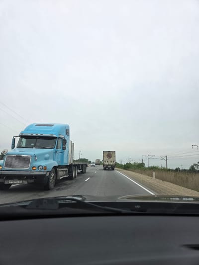 The image shows a highway scene in Ahr, Russia. A light blue Freightliner semi-truck is visible in the foreground, traveling in the same direction as the camera. In the distance, other trucks are also traveling along the highway. On the right side of the road, a barrier and vegetation are visible, with power lines extending above. The sky is overcast. The image was likely taken from inside a vehicle. The license plate of the blue semi-truck reads "M959TK 27".