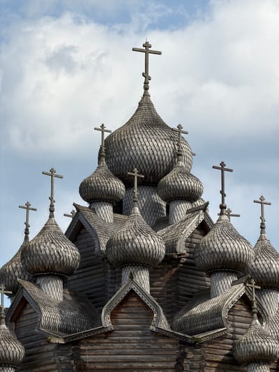 The image shows the wooden domes and crosses of the Church of the Transfiguration in Nevskii Parkleskhoz, Russia. The church, constructed in the 18th century, features a complex architectural design with multiple bulbous domes covered in wooden shingles. Several crosses of varying sizes are prominently displayed atop the domes, silhouetted against a cloudy sky. The log construction of the church's lower levels is also visible. The scene is devoid of any people, focusing solely on the intricate details of the religious structure.