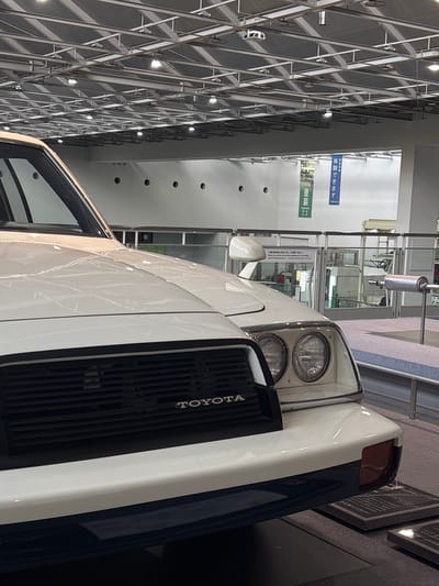 The image shows a close-up of the front of a white Toyota car on display. The car has a black grille with the "TOYOTA" logo visible. The headlights are round and dual. A dark blue bumper is visible below the white body. In the background, there are metal beams and lights on the ceiling of what appears to be an indoor exhibition hall. Two vertical banners hang in the background; one is green and reads "PAINTING" and the other is blue and reads "Experience." There is also a metal railing and a display of what looks like car parts, including an exhaust pipe. The scene appears to be a museum or exhibition, potentially related to automotive history. The exact location in Nagoya, Japan cannot be determined from the image alone, but the presence of Japanese text on the banners and the style of the car suggest it is likely in Japan.