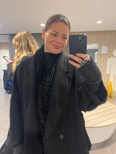 A woman in a black coat and sweater takes a selfie with a smartphone. She wears multiple rings on her left hand, which is holding the phone. Her nails are painted a metallic silver. The coat has a fuzzy trim at the neck. Another person with blonde hair is partially visible in the background, also holding a phone. The surroundings appear to be an indoor public space with light-colored walls and a bench in the background.