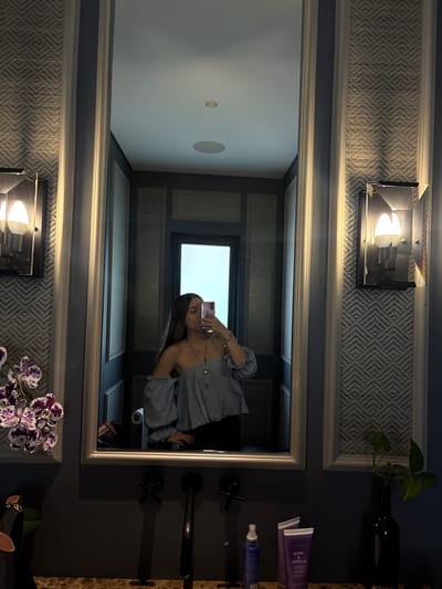 The media file is a vertical, indoor photograph taken with a smartphone, depicting a woman taking a mirror selfie. The woman, reflected in a large, ornate mirror, is wearing a blue and white striped off-the-shoulder top. She is holding a pink smartphone, capturing her reflection. Her hair is long and dark, falling over her shoulders. Her left hand is visible, adorned with a bracelet.
The setting appears to be a well-decorated bathroom or dressing area. Behind the mirror, the wall is covered in a textured wallpaper with a geometric pattern in shades of gray. A modern light fixture with a metallic frame and a bright bulb is mounted on the wall above the mirror. To the right of the mirror, a dark vanity surface is visible, holding a few toiletries, including two purple tubes of what appears to be lotion or cream with "MARY KAY BERRY & VANILLA" text and a blue spray bottle. A dark vase with a green plant is also on the vanity.
In the reflection, a window with a bright sky is visible, suggesting it is daytime. The mirror also reflects a part of the ceiling, which has recessed lighting. On the wall to the left of the mirror, another light fixture and a framed geometric pattern are visible, mirroring the details above and to the right of the main mirror. The overall impression is one of a stylish and contemporary space. The woman's pose and the selfie suggest a casual, personal moment.