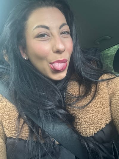 A young woman is in the driver's seat of a car, possibly in Warrington, UK, though the exact location is not evident from the image. She is wearing a seatbelt and a tan, textured jacket over a black garment. Her dark hair is styled loosely around her face. She has pierced ears, a nose ring, and is sticking her tongue out. The interior of the car is visible, with the car door and possibly a part of the exterior visible through the window.