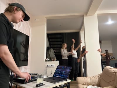 An indoor scene within a residential or event space in the city of Заозерное Снт, Russia, features a casual gathering. In the foreground left, a male subject wearing a dark cap and black t-shirt, with headphones around his neck, operates a Traktor-branded DJ controller. His right hand rests on the controller, positioned on a light-colored table alongside a MacBook laptop displaying DJ software, a glass containing a clear liquid with ice, and a black cylindrical audio adapter. A large wall-mounted television behind him reflects the silhouette of a person's raised arms. To the midground right, a group of four individuals is partially visible around a light-colored structural column or wall. One female in a light top and dark pants has her arms outstretched to her sides. Another female, in a white top and dark pants, has both arms raised above her head. A male and another female are partially obscured by the column, with their hands gripping its edges. Further in the background right, a fifth female figure in a brown outfit is partially visible in what appears to be a kitchen or dining area, with kitchen appliances visible. The space is illuminated by artificial light, revealing light-colored walls and a dark staircase in the background left. A light-colored upholstered armchair is partially visible in the foreground right.