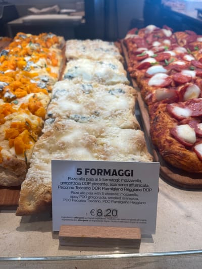 This image captures a vibrant display of various rectangular pizzas or focaccia bread, likely presented for sale in a food counter or a bakery/deli in Dorno, Italy. The pizzas are arranged on wooden serving boards, creating an appetizing visual.
Three distinct types of pizza are visible. At the top, a pizza is generously topped with slices of pepperoni or salami and white cheese chunks, resting on a golden-brown, thick crust. Below it, a "5 FORMAGGI" (Five Cheese) pizza features a creamy white cheese topping with visible blue veining, indicative of gorgonzola, alongside other white cheeses. At the bottom, another pizza showcases an orange-colored topping, possibly roasted butternut squash or sweet potato, garnished with green herbs.
A white information card is prominently displayed in front of the five-cheese pizza. The card, written in both Italian and English, specifies the ingredients for the "5 FORMAGGI" pizza: "mozzarella, spicy PDO gorgonzola, smoked scamorza, PDO Pecorino Toscano, PDO Parmigiano Reggiano." The price for this item is clearly marked as €8.20. The card also notes that ingredients and allergens are available on separate signs, and the product is made from a frozen base.
The scene suggests an indoor commercial setting during business hours, with the prepared food ready for purchase. No individuals are visible in the frame, and the time of day is not ascertainable. The close-up perspective emphasizes the textures and toppings of the different pizza varieties.