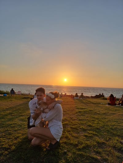 In the city of Palmachim, Israel, at dusk, a couple kneels on a grassy hillside overlooking the Mediterranean Sea. The woman, adorned with a flower crown, embraces a small dog. The man, with his arm around her, also holds the dog. In the background, the sun sets over the water, casting a warm glow. Several other individuals are scattered on the grass, some seated, appearing to enjoy the sunset. The environment is characterized by a clear sky transitioning from blue to orange, a calm sea, and lush green grass.