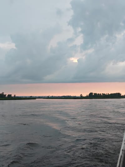 A vertical, wide-angle shot captures a serene river scene at dusk in Kazachinskoe, Russia. The sky is dominated by thick, grey clouds, with a sliver of peachy light peeking through in the upper right quadrant. A soft pink hue colors the horizon line, suggesting the last rays of sunset. Below this, a distant shoreline is visible, dotted with trees and what appear to be structures of a small settlement, possibly houses and a church spire, indicated by faint lights. The foreground is a vast expanse of dark, rippling water, with gentle waves creating a textured surface. The far right edge of the frame shows a sliver of a white structure, possibly the railing of a boat from which the photo was taken. No people or animals are visible in the frame. The overall mood is tranquil and atmospheric, characteristic of a twilight landscape.