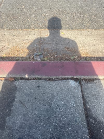 The image depicts an outdoor ground-level scene in Long Beach, United States of America, under bright sunlight. The upper portion of the frame shows a section of asphalt roadway. Below this, a strip of unkempt ground with dried leaves, twigs, and a crumpled clear plastic bag separates the road from a red-painted concrete curb. Below the curb is a section of concrete sidewalk, which is partially cracked and also contains small debris. A prominent dark shadow of a human figure, appearing to be the photographer, is cast across the asphalt, the debris strip, and the red curb. The shadow shows the head and upper torso of the individual, positioned as if facing away from the camera. The sunlight originates from the left and slightly behind the viewer, casting the shadow towards the right.