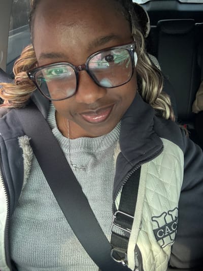 The image displays a close-up selfie of an adult female taken inside the passenger compartment of a vehicle. The specific location within Huanggu District, China, cannot be determined from the visual evidence presented.

The individual, appearing to be of African descent, has a dark complexion and is looking directly at the camera with a slight smile. She wears thick-framed, dark-colored prescription glasses, which exhibit a light-colored reflective glare, predominantly in the left lens (viewer's left). Her hair, styled with light-colored highlights or streaks, is partially visible around her face.

She is dressed in a grey, ribbed knit sweater, over which she wears a dark-colored hooded jacket. The jacket features an off-white or cream-colored quilted panel on the right side of her chest (viewer's right), bearing a dark embroidered logo or text that appears to read "CAC" with additional stylized elements above it. A silver-colored necklace with a small, stylized charm, resembling a fish or a double-curved design, is visible around her neck. A dark-colored seatbelt diagonally traverses her torso from her left shoulder to her right hip, indicating she is secured in the vehicle.

The background reveals dark-colored upholstery of vehicle seats, including headrests, consistent with the interior of a car. No other individuals or specific external environmental details are identifiable in the frame. The action captured is a self-portrait.