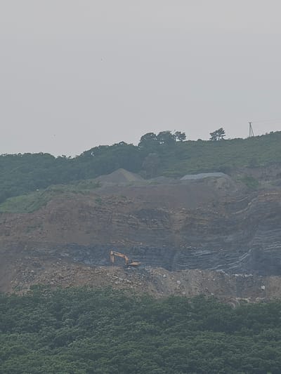 The image shows a large-scale excavation site on a hillside, with distinct layers of rock and soil exposed. A yellow excavator is visible working on one of the excavated levels. The top of the hill is covered with lush green vegetation and trees. In the distance, a power line pole with wires is visible against a hazy sky. This landscape is consistent with an industrial or mining operation. While the specific location within Bolshoy Kamen, Russia cannot be definitively determined from the image alone, such sites are often found on the outskirts of urban areas or in proximity to resource extraction activities.