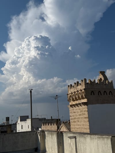 The image shows a rooftop view in Ugento, Italy, with a dramatic sky filled with large, fluffy white clouds against a bright blue sky. The sun is high in the sky, suggesting it is daytime, likely midday or early afternoon, given the bright illumination and contrast. The weather appears to be fair, with the impressive cloud formations indicating a dynamic atmosphere, possibly leading up to or following a period of rain.
Dominating the right side of the image is a portion of a building with weathered, textured walls. This building features a distinctive decorative element at the top, resembling crenellations or battlements, and a series of arched openings. Below this, other rooftops and buildings are visible, some with windows and what appear to be satellite dishes or antennas. The architecture suggests an older urban or town setting.
The scene is devoid of people. The activity taking place is simply the visual presence of the urban landscape under a striking sky. Notable details include the strong shadows cast by the buildings and structures, emphasizing the brightness of the day. The textures of the buildings, from the rough stone-like material of the prominent structure to the smoother concrete of other rooftops, are clearly visible. The overall mood is one of serene observation, capturing a moment of natural grandeur juxtaposed with human construction. There is no visible text in the image.