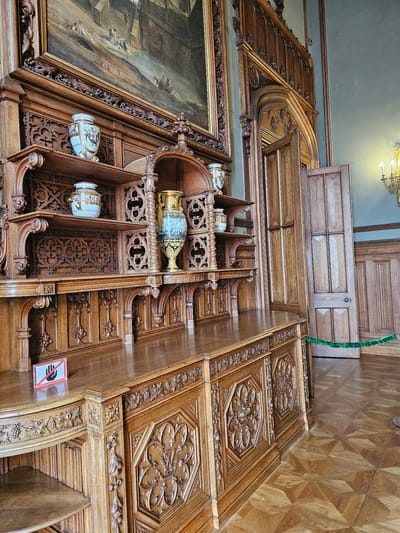 Captured from a low-angle perspective, the image showcases a segment of an ornately designed interior, presumably within the city of Alupkinskiy gorodskoy sovet, Ukraine. Predominantly crafted from wood, the scene features a richly carved wall structure, adorned with intricate patterns and floral motifs. The wall incorporates shelves that house decorative vases. A large painting with a gilded frame is visible to the left. Above, a wooden ceiling panel and a glimpse of a patterned wooden floor add to the scene's opulence. A 'Do Not Touch' sign is affixed to the wooden structure. The upper area of the photograph captures elements of the building's architecture, including a window and a lighting fixture.