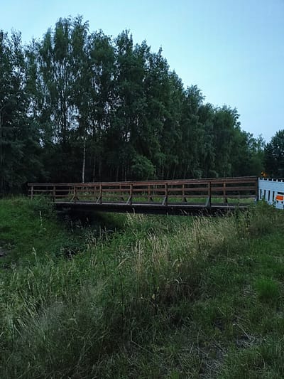 The image depicts a wooden pedestrian bridge spanning a small waterway or ditch, surrounded by dense green foliage and trees. The sky is a pale blue, suggesting either early morning or late evening. On the far right side of the bridge, a white barrier with orange accents is visible, indicating some form of construction or temporary obstruction. The scene appears to be located in a natural or semi-natural area, possibly a park or a pathway leading through undeveloped land. The overall impression is one of a quiet, outdoor setting.