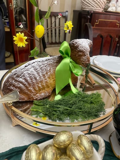 A high-angle shot depicts a mirrored gold-rimmed serving tray with various items on top of a table covered with a white tablecloth, likely indoors.
The tray holds a sliced loaf of cake dusted with powdered sugar and tied with a green ribbon, a bunch of fresh dill, and cinnamon sticks. A decorative branch with yellow flowers and a yellow egg-shaped ornament is also present. Near the edge of the tray is a portion of a white dish filled with golden-colored eggs.
The background includes a dark wooden cabinet with golden accents and some white plates. The overall scene suggests a festive occasion, possibly Easter, with a decorative and culinary display. The location in the city of Akhalkalaki, Georgia, is not evident within the visual information available in the image.