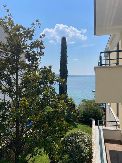 The image depicts a sunny day with a bright blue sky and scattered white clouds, overlooking a serene body of water, likely the Adriatic Sea, given the location context of Crikvenica, Croatia. The scene is viewed from a slightly elevated perspective, suggesting a balcony or upper floor of a building.
In the foreground, lush green foliage from a large tree with broad leaves dominates the lower portion of the frame, with its branches reaching upwards towards the sky. Behind the tree, a tall, slender cypress tree stands prominently. The middle ground reveals a well-maintained green lawn adjacent to a building with light-colored walls and a balcony with a green railing. A glimpse of the calm, blue water is visible beyond the garden area.
Further out, the vast expanse of the sea stretches towards the horizon, where a faint outline of distant land can be seen. A small, dark red boat is visible on the water, adding a touch of color to the tranquil seascape.
The overall impression is one of a peaceful, warm day in a coastal setting. The bright sunlight, clear sky, and calm water suggest a pleasant time of day, possibly late morning or early afternoon. There are no people clearly visible, and no text is present in the image. The scene evokes a sense of relaxation and natural beauty.