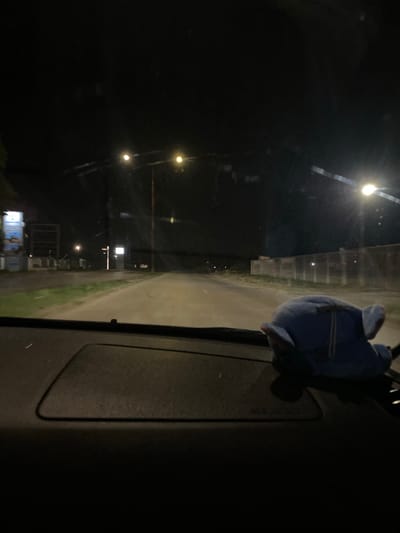 The image depicts a dark, nighttime view from inside a vehicle, looking out through the windshield. In the foreground, on the dashboard, a blue plush toy resembling an elephant is visible, resting on its side. The road ahead is illuminated by streetlights, with some vegetation visible on the left side of the road. Distant buildings and structures are faintly discernible in the background. The scene suggests a drive through a somewhat industrial or developing area at night. The specific location within the city of Yasen, Bulgaria cannot be determined from the visual information provided.