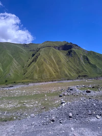 The image shows a wide shot of a mountainous landscape under a bright blue sky with scattered white clouds. The foreground is a rough, rocky terrain covered with sparse grass. A low-lying area with more dense grass and some scattered rocks separates the foreground from the base of a large, green mountain. The mountain slopes are covered in lush green grass, with visible undulations and some darker patches, possibly indicating vegetation or shadows. A steep, rocky cliff face is visible towards the top of the mountain. The scene appears to be an outdoor natural environment. The image does not provide any specific details or context that would allow for identification of the location in Almasiani, Georgia, or any people, subjects, objects, actions, or interactions.