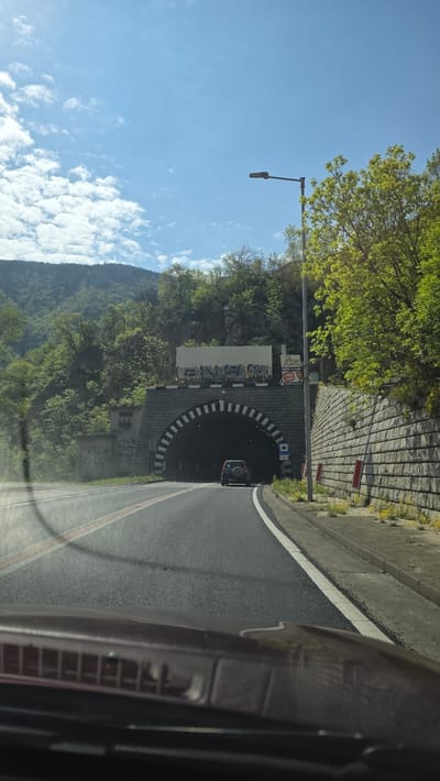 The image captures the view from inside a car, seemingly approaching the entrance of a tunnel located in Stara Kresna, Bulgaria. The road leading to the tunnel is paved and marked with lane dividers. The tunnel's entrance is characterized by alternating black and white stripes forming an arch. To the left of the tunnel entrance, a mountain covered with green trees and vegetation is visible. The sky above is blue with scattered white clouds. A street lamp stands nearby. A car is emerging from the tunnel. The right side of the image is dominated by the interior of the vehicle from which the image is taken.