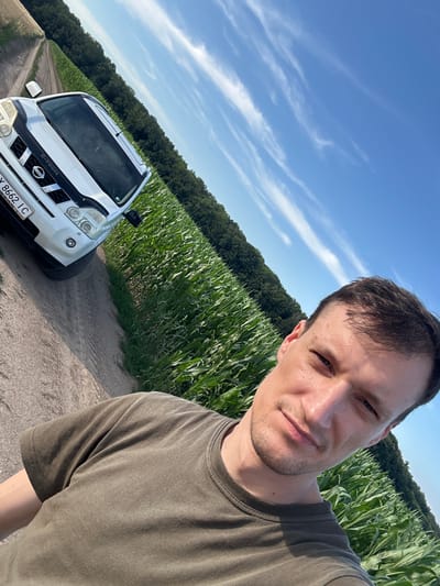 A man takes a selfie outdoors next to a white Nissan X-Trail. The car is parked on a dirt road beside a tall green cornfield. A dense forest borders the field, and the sky above is a clear blue with thin white clouds. The man is wearing a dark olive t-shirt. The license plate on the car reads "YX 8662 IC". The scene is likely in or near the village of Slobidka-Zalisetska, Ukraine.