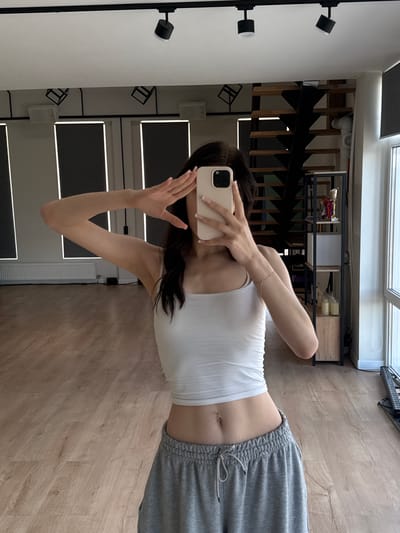 The media file shows a person taking a mirror selfie in what appears to be a modern apartment or studio in Pokrovskoe, Russia. The person is wearing a white tank top and grey sweatpants. They are holding a smartphone with a light pink case in their right hand, with their left hand slightly raised. Their reflection is visible in a large mirror that covers a significant portion of the wall.
In the background, seen through the mirror, is a staircase with black railings and wooden steps, suggesting an interior space with multiple levels. There are shelves mounted on the wall above the staircase, displaying various objects, including what look like decorative items and bottles. The overall aesthetic suggests a contemporary design.
The lighting in the room appears to be artificial, with track lighting visible on the left side of the frame and light strips integrated into the wall panels behind the person. The floor is made of light-colored wood planks.
The image is oriented horizontally, and the person's body is mostly visible from the torso down. The focus is on the person and their outfit, as well as the surrounding interior elements. There is no visible text in the image. The time of day is not discernible, and the weather is irrelevant to the interior setting. The overall mood is casual and relaxed, typical of a personal selfie.