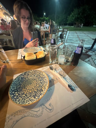The image captures a dimly lit outdoor dining scene at a restaurant in Asprovalta, Greece. The focus is on a wooden table laden with dinnerware and drinks. In the foreground, a woman wearing glasses and a dark jacket over a blue and yellow patterned dress holds a smartphone, her fingers poised as if texting or browsing. Beside her, a small black oval dish contains two golden-brown bread rolls. Two patterned ceramic plates, one with a blue and white intricate design, are placed on the table, along with several glasses, a bottle of Pepsi, and cutlery. In the background, other diners are visible at their tables, and the setting suggests a warm evening with trees and a paved area visible. The overall atmosphere appears relaxed and social, typical of a restaurant setting in a Greek coastal town.