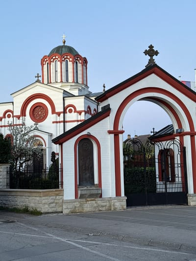 The image shows an Orthodox church in Trstenik Municipality, Serbia. The building is white with red accents around windows, arches, and roof edges. A black metal fence surrounds the property. The sky is clear and blue. There are no people visible in the image.