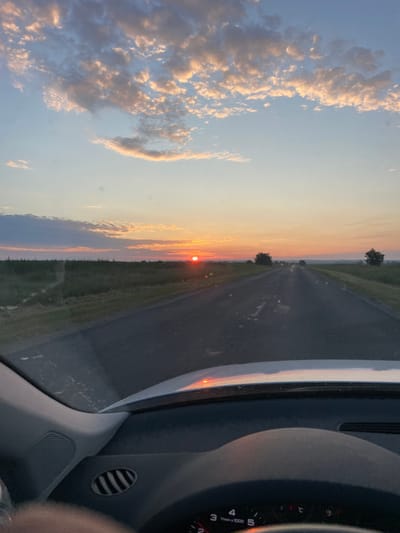 The image captures a view from inside a car driving on a road at sunset. The sun, a bright orange orb, is low on the horizon, casting a warm glow across the sky. Clouds, tinged with pink and gold, are scattered across the blue expanse. The road ahead stretches into the distance, flanked by green fields. The interior of the car is visible in the foreground, including a dashboard with a speedometer. The scene is set in Onițcani, Moldova.
