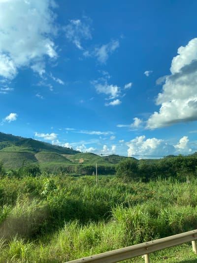 The image depicts a rural landscape with rolling hills covered in lush green vegetation under a bright blue sky dotted with white clouds. The foreground features tall grasses and some trees, with a grey metal railing visible at the bottom. In the middle ground, more trees and the lower slopes of the hills are seen, with evidence of terracing on some of the hillsides. A single utility pole stands amidst the greenery. The background shows the further expanse of the hills, receding into the distance. The scene is peaceful and natural, likely captured from a vehicle due to the angle of the railing. The location is identified as Thôn 18, Vietnam, which is consistent with the tropical or subtropical vegetation and hilly terrain.