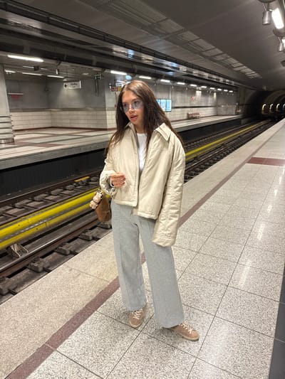 A person stands in a metro station. The person is wearing light grey sweatpants, a white shirt, a tan bomber jacket, and tan shoes. The person is also wearing sunglasses. The station has grey and brown tiled floors and a dark ceiling with lights. The train tracks are visible.
