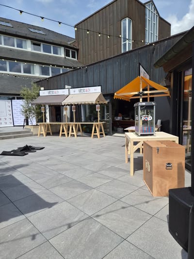 The scene depicts an outdoor commercial area in Lenningen, Luxembourg. There are two outdoor pop-up shops or stands with canopies, branded "LET'Z POWERED BY." The first one has an orange canopy, the second a beige one. A clear glass display case with colorful items is visible in one stand. The stands are constructed of light-colored wood. Nearby, there is a wooden box with a handle and an image of a monkey on it. A speaker is also visible in the shot. The foreground is paved with gray, square tiles. The background consists of buildings with black and brown exterior elements. There are windows and lights strung along the building. There are also "MERCURE" posters on the front. The sky with some clouds is partially visible in the upper-left corner. A tree is visible between the "LET'Z POWERED BY" signs.