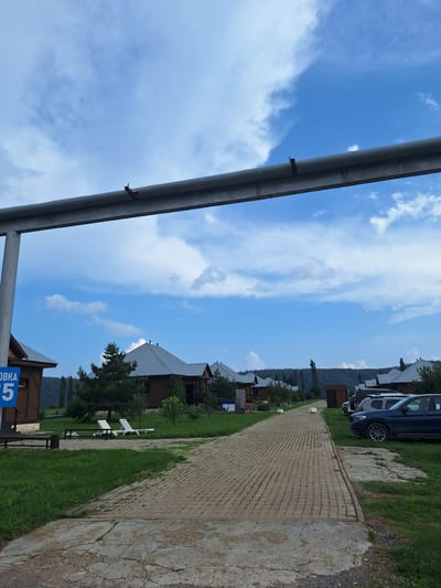 The image depicts a paved pathway leading through a village-like settlement with wooden houses featuring gray roofs. The pathway is lined with green grass and scattered trees. On the left side of the path, there are two white lounge chairs on the grass, and a blue sign with white Cyrillic lettering and the number "5". Several cars are parked on the right side of the path. In the background, a dense forest covers the rolling hills under a blue sky with scattered white clouds. A large metal pipe runs horizontally across the upper portion of the image, supported by vertical structures. The overall scene suggests a peaceful, rural setting. The location is identified as Atamanovka, Russia.