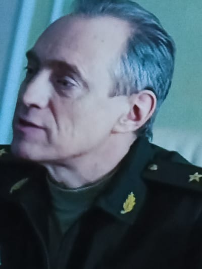 A close-up, head-and-shoulders shot shows a middle-aged Caucasian man with receding gray hair and a pale complexion, seen from the left profile. He wears a dark military uniform with gold epaulets, one featuring a star and another a leaf-like emblem. The man's mouth is slightly open, suggesting he is speaking. His facial expression is difficult to determine due to the image quality and angle, but appears neutral. The background consists of a gradient of soft blue and green hues, reminiscent of a plain studio backdrop or a softly lit wall. The lighting appears diffused and even, with no harsh shadows. There is no visible indication of the specific location within Gusinoozyorsk, Russia.