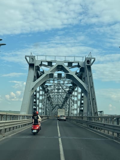 The image depicts a view from a vehicle traveling on a bridge, likely the Giurgiu–Ruse Friendship Bridge, which connects Giurgiu, Romania, to Ruse, Bulgaria. The bridge is a large, gray, steel truss structure. The road is paved and has lane markings. In the foreground, a red scooter is traveling in the same direction as the vehicle, carrying two individuals. The person in the back is wearing a black t-shirt with a white logo. The scooter has a license plate that reads "GR 53 WMW". Further ahead on the bridge, a silver car and a white truck with "Glo-Cargo" written on it are visible. The sky is blue with scattered white clouds. The environment appears to be open country with some vegetation visible in the distance.