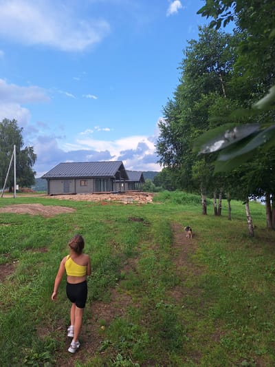 In Atamanovka, Russia, on a partially constructed property with green grass and dirt paths, a young girl with a ponytail, wearing a yellow one-shoulder top and black shorts, walks away from the camera. She is followed by a medium-sized dog with brown and black fur, walking on a dirt path. In the background, several modern, grey, single-story houses with dark roofs are visible. Tall green trees, including birches, line the right side of the frame. The sky is a mix of blue with scattered white clouds.