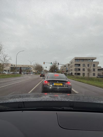 The image depicts a view from inside a car, looking forward onto a rainy city street in Bertrange, Luxembourg. A black car is in front with a yellow license plate reading "UU 5052". There are traffic lights overhead. On the sides of the road are buildings, with some showing modern architecture. Street lamps line the curb. The sky is overcast.
