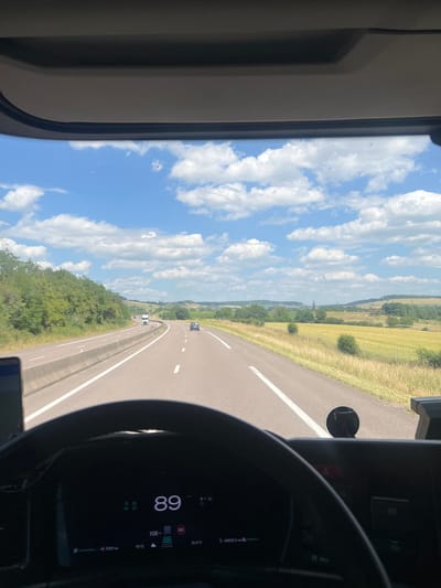 This media file shows a view from the driver's seat of a vehicle, likely a truck, as it travels on a highway in Rolampont, France. The scene is a daytime shot with a bright, blue sky filled with scattered white clouds. The road ahead curves to the right, bordered by green trees and fields of golden crops.

The driver's side dashboard is visible on the right, featuring a digital display. The display shows a speed of 89 km/h, an external temperature of 31.4°C, and the time as 15:12. There is also a speed limit indicator showing 80. Other indicators and text are present but not entirely legible. A steering wheel is partially visible in the foreground. The overall impression is of a journey on a sunny day, possibly during summer, with clear visibility and fair weather. The image captures the perspective of someone driving through a rural French landscape.