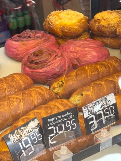 The image shows a display of baked goods, likely in a bakery or market, in the city of Stryi, Ukraine. In the foreground, there are several golden-brown croissants or similar pastries. Behind them, two long, braided pastries with visible filling are displayed. Further back, there are pink-frosted swirl pastries and some braided buns topped with crumbs. Two price tags are visible in the foreground. One tag has handwritten text and the number "19.95" displayed digitally. The other tag has handwritten text and the number "37.90" displayed digitally. The environment appears to be an indoor setting with a shelf or display case.