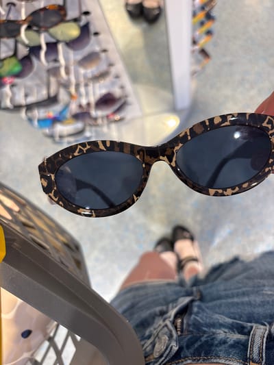 A person's hand is holding up a pair of leopard print sunglasses. The sunglasses have dark lenses and a thick, patterned frame. In the background, a display of sunglasses is visible on a rack. The person is wearing denim shorts and sandals. The scene appears to be inside a store, possibly an optical shop or a department store, given the display of merchandise. The flooring is light-colored and patterned. The lighting suggests it is daytime.