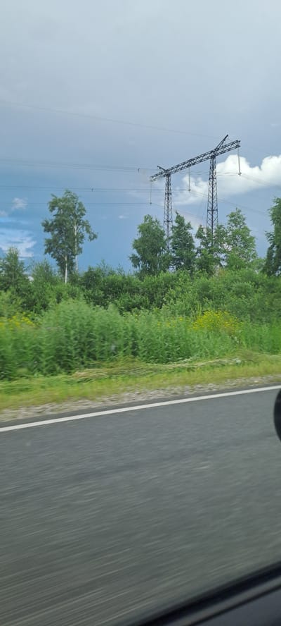 A scene captured from a moving vehicle shows a landscape with a prominent power line structure against a cloudy sky. The image is dominated by lush green vegetation, including trees and tall grass, with some yellow wildflowers interspersed. A paved road with a white line marking its edge runs horizontally across the lower part of the frame, indicating motion blur consistent with a moving perspective. In the background, a large, dark metal power line pylon with cables stretching across the sky is visible. The sky itself is a mix of light blue and gray clouds, suggesting possibly overcast or pre-storm conditions. The location is identified as Ульянов ауылдық округі, Kazakhstan.