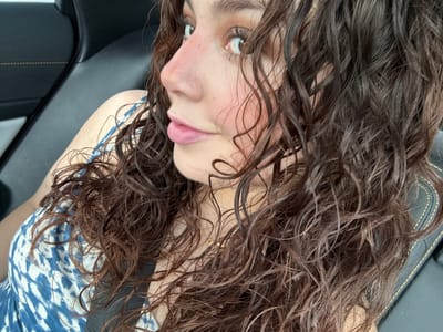 A woman with damp, dark curly hair is angled upside down, possibly in a car. She's wearing a blue and white patterned top and a seatbelt. Her face is visible, showing her eyes, nose, and lips. The background includes a black car interior, suggesting a relaxed or candid moment.