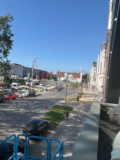 The image depicts a street scene in Molenbeek-Saint-Jean, Belgium, characterized by ongoing construction work. A street is lined with parked cars, including a red sedan, a white van, and a black SUV, all on a cobblestone surface. The construction zone is demarcated by yellow and blue barriers, with visible stacks of construction pipes and a large green water tank in the background. Several construction vehicles, including a concrete pump, are present. A white truck is parked on the left side of the street.
In the mid-ground, a paved sidewalk runs alongside the construction. A few individuals are walking on the sidewalk, and a person is riding a bicycle. Buildings of varying architectural styles line the street, with some featuring graffiti. The sky is clear and blue, indicating a sunny day. The perspective suggests the photo was taken from a slightly elevated position, possibly from a building overlooking the street. Shadows from trees and buildings are cast across the ground.