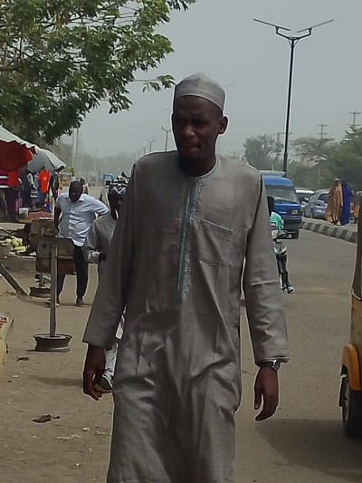 This image captures a daytime scene on a street in Daurawa, Nigeria, under a hazy sky.
In the foreground, a dark-skinned male, estimated to be an adult, walks directly towards the viewer, slightly angled to the right. He wears a light grey traditional tunic (kaftan) featuring light blue embroidery along the central placket and a single patch pocket on the left chest. A light grey skullcap (kufi) covers his head. A dark-colored wristwatch is visible on his left wrist. His expression is neutral.
To the immediate left of the foreground subject, market activity is visible. Several stalls with various goods, including green, spherical produce resembling watermelons or gourds, are set up beneath red and grey tarps or umbrellas. Another dark-skinned male, wearing a light blue collared shirt and dark trousers, stands behind a market stall, facing right. Further left, a metal stand, possibly part of a weighing scale, is on the unpaved, dusty ground.
In the background, a paved road stretches into the distance on the right side of the frame. Traffic includes a prominent blue van or minibus moving away from the viewer, and other vehicles are visible further down the road. On the right shoulder of the road, two individuals, clad in full-body coverings (one brown, one blue), walk away from the camera. Modern streetlights with flat-panel fixtures are positioned along the road. A yellow vehicle, possibly an auto-rickshaw, is partially visible on the extreme right foreground. Lush green foliage from trees is visible in the upper left and scattered throughout the background. The sky is uniformly light grey, indicating overcast or hazy conditions.