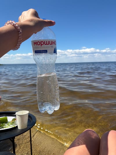 In Lebedivka, Ukraine, a hand adorned with a pink beaded bracelet holds a partially full plastic bottle of "МОРШИНСЬКА" natural mineral water. The bottle, containing clear liquid and visible condensation, is suspended against a bright blue sky with scattered white clouds. The backdrop is a body of calm water, its surface reflecting sunlight, suggesting an outdoor setting, possibly near a beach or lakeside. In the lower left corner, a small black table holds a white paper cup and a plate with what appears to be food items, including a green vegetable and herbs. The sandy shore is visible in the foreground, where the knees of a person are also present. The overall scene depicts a relaxed moment outdoors, with refreshments and a clear sky.