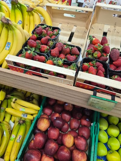 The image depicts a static produce display inside a retail environment, located in the Municipal Unit of Corinth, Greece. The upper left quadrant features multiple bunches of ripe yellow bananas, several bearing blue Dole brand stickers. To the right of the bananas, numerous black and clear plastic punnets are filled with red strawberries, many with green caps and stems, arranged within and upon light-colored wooden crates. A wooden shelf structure divides the upper and lower display tiers. The lower left section displays additional bunches of ripe yellow bananas in a green plastic crate, also with blue stickers. Adjacent to these, in the center, a green plastic crate contains a large quantity of red apples with varying hues of red and some yellow areas. To the far right of the lower section, another green plastic crate is filled with green apples. A green-edged price label with white text, partly obscured, is visible above the red apples, displaying "ΓΕΝΙΚΟΙ ΚΑΝΟΝΕΣ ΠΑΡΑΓΩΓΗΣ" (General Production Rules) and a price, likely 2.50€. No individuals are present in the scene.