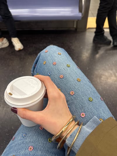 The image depicts an interior view of a public transit vehicle, identified as being in Hoboken, United States of America. The primary focus is the upper legs and right arm of a seated individual. This individual wears light blue denim jeans extensively adorned with small, multi-colored flower-shaped beads or appliqués. Their right hand, with dark red/burgundy fingernail polish, holds a white disposable coffee cup with a white "SOLO Traveler lid." Multiple gold or bronze bangles are visible on the wrist of the holding arm, and the cuff of a dark green or khaki sleeve is also present.
The floor of the vehicle is dark grey. In the background, to the left, the lower legs of another person wearing dark pants and white sneakers are partially visible. To the right, the lower legs and black shoes of a third individual, also in dark pants, are visible, standing near a yellow safety strip or line on the floor. A light purple/blue bench seat or railing is present in the mid-background.