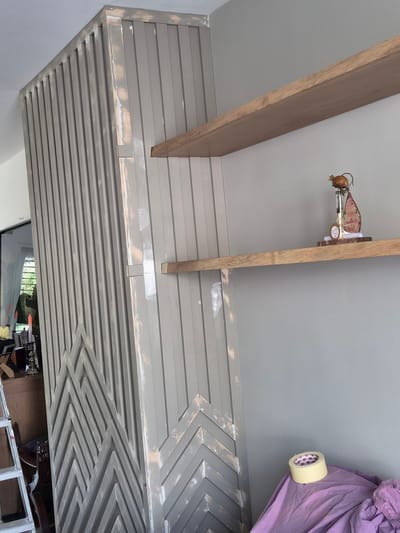 The image depicts an interior scene, likely a room undergoing renovation or decoration. A tall, fluted partition, painted a muted grey, dominates the left side of the frame. It features geometric patterns at its base, resembling inverted chevrons or zigzags. To the right, two light-colored wooden shelves are mounted on a light grey wall. The top shelf holds a small decorative item, possibly a figurine with a straw-like element on its head and a pink flag bearing the inscription "Happy Mother's Day". A roll of masking tape sits on a draped purple cloth on the lower shelf. In the background, through a glass door or window, a glimpse of an outdoor area with greenery is visible, along with part of a ladder. The overall impression is one of a domestic space in a state of flux. The location in Schengen, Luxembourg, cannot be determined from the visual information provided.