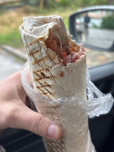 A person is holding a large doner kebab wrapped in plastic. The kebab is filled with meat, tomatoes, some kind of shredded ham-like meat, and potatoes, all wrapped in a grilled tortilla. The person appears to be in a car. The car's side mirror is visible and shows a blurred cityscape. Based on the reflections in the car mirror and available background details, it is likely the city of Enem, Russia.