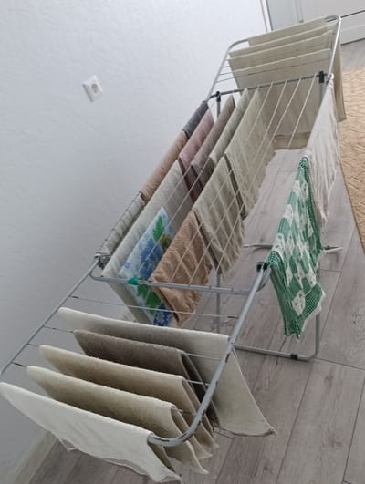 This is a medium-angle, eye-level shot of a metal clothes drying rack filled with various towels and cloths. The drying rack is positioned on a light gray wooden floor against a white textured wall. The wall has a single white electrical outlet visible. The towels and cloths are of different colors and patterns, including light beige, grey, pink, white with blue floral patterns, and a green and white checkered cloth. Some of the cloths are folded, while others are hanging loosely. The lighting is bright and even, suggesting an indoor environment. The scene appears to be a domestic setting in Marshyntsi, Ukraine, likely a laundry room or bathroom.