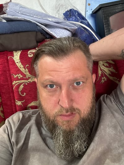 The image shows a close-up, overhead view of a man lying on a red couch with a gold ornate pattern. The man has a full grey beard and blue eyes, and he is looking directly at the camera. His right arm is behind his head, and he is wearing a grey t-shirt. In the background, there are stacks of folded clothing, some of which are in clear plastic packaging. To the right of the man's head is a black object that resembles a miniature fireplace. The overall scene appears to be an indoor setting, likely a home environment in Schokino, Russia, as suggested by the context provided.