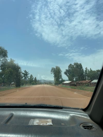 The image, taken from inside a vehicle, depicts a dirt road in a rural environment. On the left, there is a blue sky with scattered white clouds. Trees and sparse vegetation line the side of the road. Some structures are visible amongst the trees. The dashboard of the vehicle is visible on the right side of the frame. The windshield has a visible water stain. It is not possible to confirm this image was taken in the city of Zankan, Nigeria, or to discern people, specific actions, or interactions within the scene.